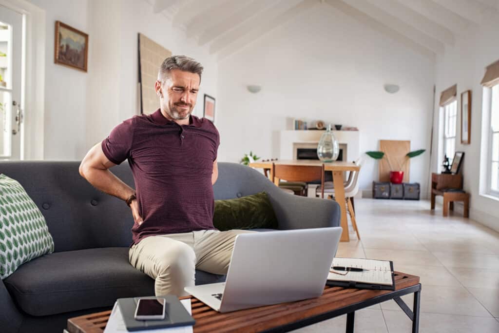 Tired mature businessman stretching back while working from home. Business man suffering from back pain while working on laptop. Stressed mid adult man teacher taking a break after long hours at work sitting on couch.